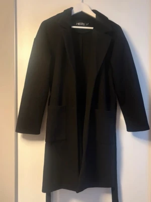 Svart kappa från Pieces - Stilren svart kappa från Pieces med raka ärmar och två stora fickor framtill. Kappan har en klassisk krage och bälte i midjan för en snygg siluett. Perfekt för dig som gillar minimalistisk och tidlös design.