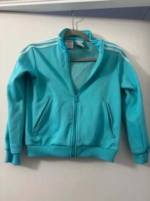 Turkos Adidas ziptröja med ränder - Snygg turkos ziptröja från Adidas med klassiska vita ränder på axlarna. Tröjan har dragkedja framtill, två fickor med dragkedja och ribbade muddar vid ärmslut och nederkant. Perfekt för en sportig och avslappnad stil.