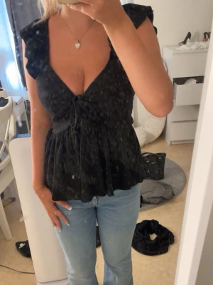 Svart topp volangdetaljer - Snygg svart omlottopp med volangdetaljer vid axlarna och v-ringning. Toppen har knyt framtill och är lätt utsvängd nedtill för en feminin siluett. Perfekt att styla med jeans eller kjol för en trendig look.