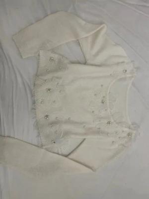 Vit stickad tröja med pärlor och blommor - Supersöt vit stickad tröja med långa ärmar, dekorerad med stora broderade blommor och pärlor på fram- och nederkant. Tröjan har rund halsringning och ribbade muddar. Perfekt för dig som gillar romantisk och unik stil.
