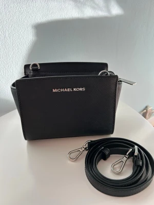 Svart axelväska från Michael Kors - Säljer en Michael Kors Selma Saffiano leather crossbody. Endast använd fåtal gånger, i väldigt  fint skick. Stilren svart axelväska  i läder med silverfärgade metalldetaljer och avtagbar axelrem. 