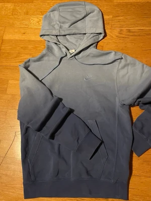 Nike hoodie i blått ombre - Snygg hoodie från Nike i en blå ombre-färg som går från ljusare upptill till mörkare nedtill. Klassisk huva och magficka framtill, samt broderad Nike-logga på bröstet. Perfekt för en avslappnad och sportig stil.