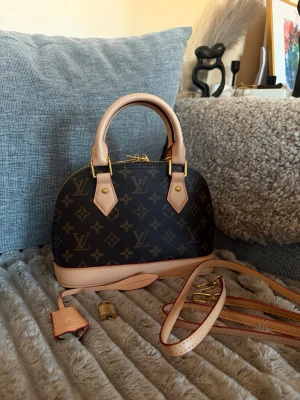 Louis Vuitton Alma BB Monogram väska - Superfin Louis Vuitton Alma BB handväska i klassiskt brunt monogramcanvas med ljusa läderdetaljer och guldfärgade metalldelar. Bågeformad modell med två handtag och avtagbar axelrem. Kommer med hänglås och nyckel. Perfekt storlek för det viktigaste.