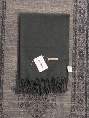 Acne Studios Halsduk - Snygg mörkgrå halsduk från Acne Studios i mjuk ull. Klassisk design med fransar i kanterna och diskret logotyp. Perfekt att svepa runt halsen för en stilren look. Passar till det mesta och är enkel att matcha med olika outfits.
