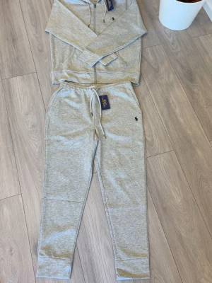 Grått loungewear-set från Polo Ralph Lauren - Mysigt loungewear-set i ljusgrått från Polo Ralph Lauren. Setet består av en hoodie med dragkedja och fickor samt matchande byxor med dragsko i midjan och muddar vid benslut. Båda delarna har den klassiska Polo-loggan broderad på bröstet och benet.