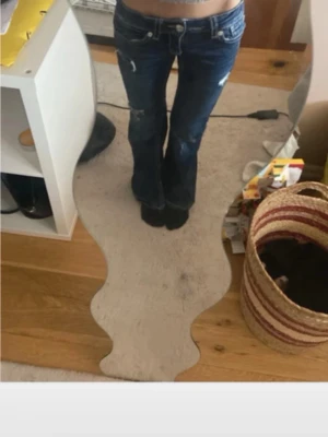 Mörkblå bootcut jeans med slitningar - Säljer ett par mörkblå bootcut jeans med slitningar på låren och snygga broderade detaljer på bakfickorna. Jeansen har låg midja och klassisk femficksdesign. Perfekta för en avslappnad och trendig look.