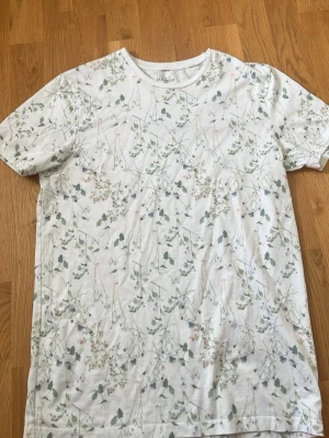 Vit blommig t-shirt Tailored Originals - Snygg vit t-shirt från Tailored Originals med ett diskret blommigt mönster i grönt och ljusa färger. Klassisk rund halsringning och korta ärmar. Perfekt för dig som gillar en stilren men ändå unik look.