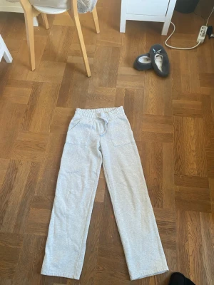 Grå mjukisbyxor med raka ben - Snygga ljusgrå mjukisbyxor med raka ben och resår i midjan samt dragsko. Byxorna har två stora fickor fram och två bakfickor. Perfekta för en chill och avslappnad stil. Tillverkade i mjukt och bekvämt material.kan bli low waiste om man viker ner midjan
