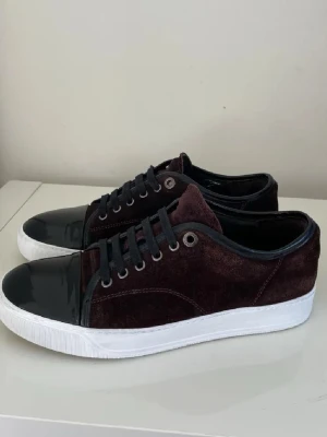 Vinröda sneakers med lackad tå - Snygga sneakers i vinröd mocka med svart lackad tå och svarta skosnören. Skorna har vit sula och metallfärgade öljetter. Perfekta för dig som vill sticka ut med en unik stil och coola materialkombinationer.