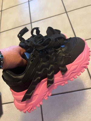 Svarta sneakers med rosa sula - Säljer ett par riktigt snygga sneakers i svart med chunky rosa sula. Skorna har grova svarta snören, meshdetaljer och coola vågiga detaljer på sidorna. Perfekta för dig som vill sticka ut med färg och streetstyle. Materialet är en mix av syntet och mesh.