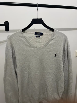 Grå v-ringad tröja från Polo Ralph Lauren - Snygg grå v-ringad tröja från Polo Ralph Lauren med klassisk broderad logga på bröstet. Tröjan har långa ärmar och ribbade muddar vid ärmslut och nederkant. Perfekt att matcha med jeans eller skjorta för en clean look.