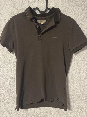 Brun pikétröja från Burberry - Snygg brun pikétröja från Burberry med klassisk krage och korta ärmar. Tröjan har knappar vid halsen och diskret Burberry-logga på bröstet. Perfekt till jeans för en stilren look. Tröjan är S men rätt liten så passar XS bättre. Kan gå ner i pris vid snabb affär!
