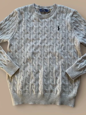 Grå kabelstickad tröja Polo Ralph Lauren - Snygg grå kabelstickad tröja från Polo Ralph Lauren med rund halsringning och den klassiska broderade loggan på bröstet. Tröjan har ribbade muddar vid ärmslut och nederkant, perfekt för en stilren och avslappnad look. Det står storlek M men passar bättre som storlek S. Modellen är 185 och väger 65