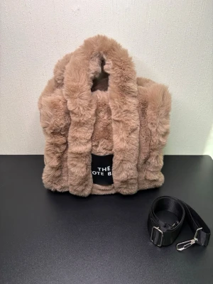 Beige fluffig handväska från Marc Jacobs - Supersöt beige handväska i mjuk fuskpäls med två handtag och avtagbar svart axelrem. Väskan har en fyrkantig form och en svart patch framtill med texten 'THE TOTE BAG'. Perfekt för dig som vill sticka ut med en mysig accessoar.