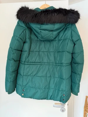 Grön dunjacka Tommy Hilfiger L - Snygg grön dunjacka från Tommy Hilfiger med huva och svart fuskpäls. Jackan har quiltad design, dragsko nertill och klassisk logga på insidan. Perfekt för kalla dagar och riktigt trendig look.