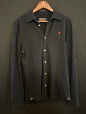  Morris skjorta  (inkl. gratis frakt) - Stilren svart skjorta från Morris med klassisk krage och vita knappar. På bröstet finns ett orange broderat emblem som ger en snygg detalj. Skjortan är långärmad och tillverkad i mjuk bomull, perfekt för en chill och clean look. 97% bomull 3% Elastan