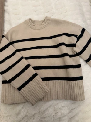 Beige stickad tröja med svarta ränder - Säljer en beige stickad tröja från H&M med svarta horisontella ränder. Tröjan har rund halsringning, långa ärmar och bred ribbstickad mudd vid ärmslut och nederkant. Perfekt för en avslappnad och trendig look.