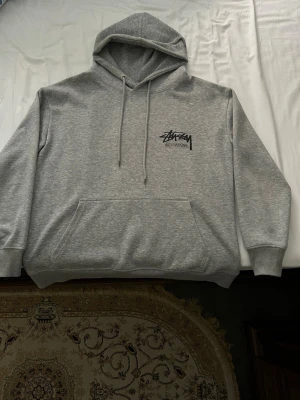 Grå Stüssy hoodie med logga - Säljer en grå hoodie från Stüssy med svart logga på bröstet och stor logga på ryggen. Klassisk känguruficka framtill, justerbar huva med dragsko och ribbade muddar. Tillverkad i mjukt sweatshirtmaterial, perfekt för en avslappnad streetwear-look.