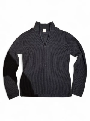 Mörkgrå stickad tröja med halv dragkedja - Mörkgrå stickad tröja med ribbad struktur och halv dragkedja framtill. Tröjan har hög krage och långa ärmar, perfekt för kyliga dagar. Materialet ser ut att vara mjukt och värmande, passar till många olika stilar.