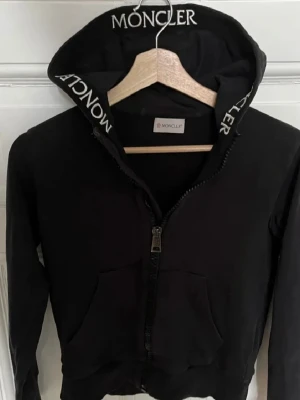 Svart hoodie från Moncler med dragkedja - Svart hoodie från Moncler med vit logga på huvan. Hoodien har dragkedja framtill, två fickor och ribbade muddar. Tillverkad i mjukt material som känns skönt mot huden. Perfekt för en avslappnad och trendig look.