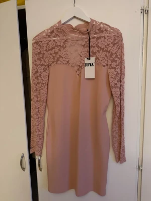 Rosa spetsklänning från Make Way - Helt ny, Supersnygg rosa kortklänning från Make Way med långärmade spetsdetaljer och hög hals. Klänningen har blommigt spetsmönster upptill och på ärmarna, samt en figurnära passform. Perfekt för dig som vill sticka ut lite extra!