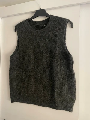 Mörkgrå stickad väst - Mysig mörkgrå stickad väst med rund halsringning och ribbad struktur. Perfekt att slänga över en skjorta eller t-shirt för en avslappnad men ändå stilren look. Västen har en rak passform och är enkel att matcha med olika outfits.