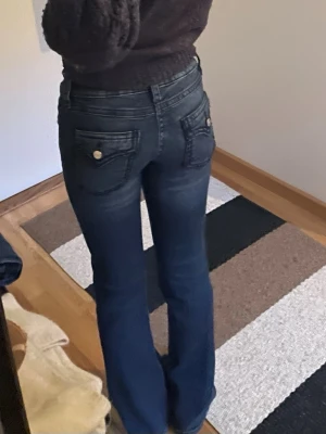 Mörkblå bootcut jeans med fickdetaljer - Snygga mörkblå bootcut jeans med dekorativa bakfickor och silverfärgade knappar. Jeansen har låg midja och klassisk femficksdesign. 