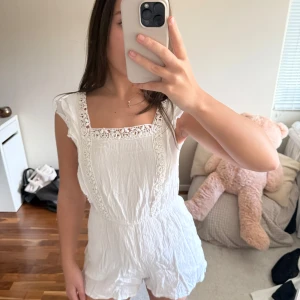 Vit spetsjumpsuit med volangdetaljer - Supersöt vit jumpsuit med spetsdetaljer och volang vid axlarna. Kort modell med knytning i ryggen och fyrkantig ringning. Tunt, krinklat tyg som känns luftigt och somrigt. Perfekt för dig som vill ha en söt och trendig look.