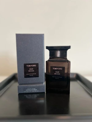 Tom Ford Oud Wood EdP 100ml - Tom Ford Oud Wood Eau de Parfum i en elegant mörkbrun glasflaska med svart etikett och matchande kartong. Flaskan rymmer 100 ml och har en lyxig, stilren design som passar perfekt på parfymhyllan. En exklusiv doft med träiga och orientaliska toner.