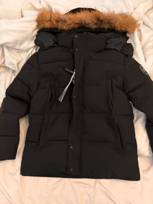 Svart dunjacka från Canada Goose - Snygg svart dunjacka från Canada Goose med avtagbar huva och pälsdetalj. Jackan har patch med logga på ärmen, dragkedja och knappar framtill samt praktiska fickor. Perfekt för kalla vinterdagar och riktigt varm tack vare dunfyllningen.