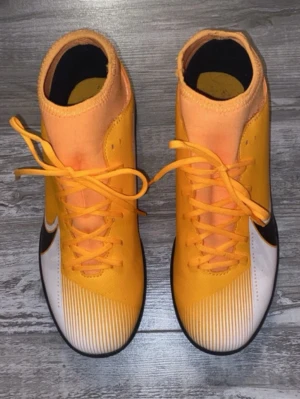 Nike Mercurial orange fotbollsskor - Säljer ett par Nike Mercurial fotbollsskor i en snygg orange färg med vita och svarta detaljer. Skorna har hög krage för extra stöd, snörning och slitstark yttersula för bra grepp på planen. Tillverkade i syntetmaterial och designade för snabbhet och kontroll.