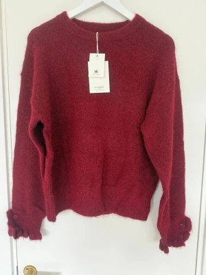 Mörkröd fluffig stickad tröja - Mysig mörkröd stickad tröja med rund hals och långa ärmar. Tröjan har dekorativa knappar på ärmsluten och är gjord i ett fluffigt material som känns extra mjukt. Perfekt för att hålla värmen under kyliga dagar.