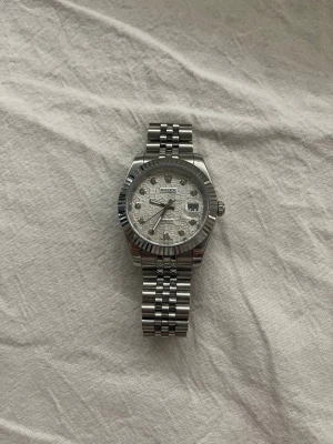 R- silverfärgad klocka - Stilren silverfärgad R- klocka med länkarmband i rostfritt stål. Urtavlan är ljus med mönster och tydliga indexmarkeringar samt datumfönster. Klassisk räfflad bezel och robust design som ger en exklusiv känsla.