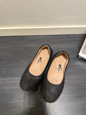 Svarta ballerinaskor med söm - Snygga svarta ballerinaskor med rund tå och dekorativ vit söm runt sulan. Skorna har en beige innersula med perforering för extra komfort. Yttermaterialet är troligtvis syntet och sulan är i gummi. Perfekta för en stilren och enkel look.