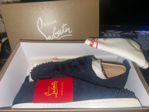Marinblå Christian Louboutin sneakers - Snygga marinblå sneakers från Christian Louboutin med klassiska röda sulor och svarta nitar på tån. Skorna är låga och har ovandel i mocka, vita yttersulor och svarta skosnören. Perfekta för dig som vill sticka ut med exklusiv design.