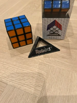 Rubiks kub 3x3 med ställ (går att köpa styck) - Rubiks kub 3x3 i klassisk design med färgerna blå, orange, gul och vit. Kuben kommer med en svart trekantig ställning och originalförpackning. Perfekt för dig som gillar kluriga pussel och vill utmana hjärnan. De är även en som är helt oanvänd och den andra är använd 1 gång. Pris kan diskuteras💕