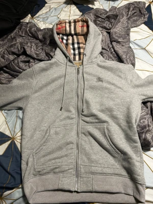 Grå zip hoodie från Burberry XL - Snygg grå zip hoodie från Burberry med klassiskt rutigt foder i huvan och diskret logga på bröstet. Hoodien har dragsko i huvan, två stora fickor fram och ribbade muddar. Perfekt för en chill och stilren look. Nypris ca 4-5 tusen men säljer denna för endast 3 tusen tröjans storlek är XL men passar mer som L