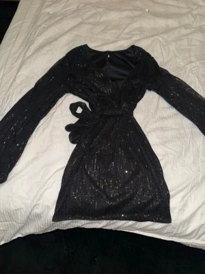 Svart glittrig omlottklänning - Snygg svart kort omlottklänning med glittriga detaljer och lång ärm. Klänningen har ett knyt i midjan och v-ringning, perfekt för att sticka ut. Materialet känns mjukt och följsamt.