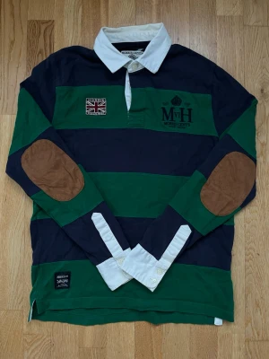 Randig rugbytröja Morris Gent's S - Klassisk långärmad rugbytröja från Morris Gent's i grönt och marinblått med breda ränder, vit krage och bruna armbågslappar. Broderade detaljer på bröstet och patch med brittisk flagga. Tillverkad i bomull för en skön och avslappnad stil.