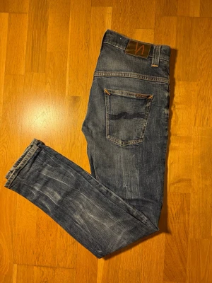Blå washed Nudie Jeans ”Tape Ted” - Snygga blå jeans från Nudie Jeans med klassisk femficksdesign och coola slitningar. Bakfickorna har Nudie Jeans ikoniska broderi och läderpatch i midjan. Jeansen är i denim med orangea sömmar.