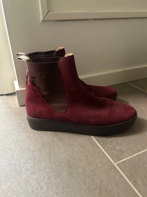 Vinröda Chelsea boots från Gant - Snygga vinröda Chelsea boots från Gant i mocka med elastiska sidopaneler och svart sula. Klassisk siluett med dragflik bak för enkel påtagning. Perfekta för dig som vill ha en stilren och trendig look.