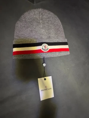 Grå Moncler mössa med ränder - Snygg grå mössa från Moncler med klassiska svarta, vita och röda ränder runt kanten. Mössan har Moncler-logga framtill och är tillverkad i mjukt stickat material. Perfekt accessoar för att lyfta din streetstyle.