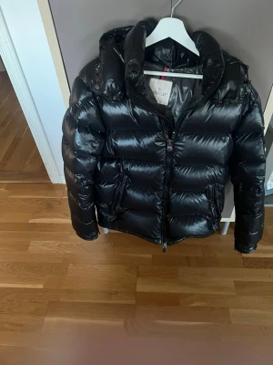 Svart pufferjacka från Moncler - Snygg svart pufferjacka från Moncler med glansig finish och stor huva. Jackan har dragkedja framtill, två sidofickor och en ficka med Moncler-logga på ärmen. Perfekt för kalla vinterdagar och riktigt trendig look.