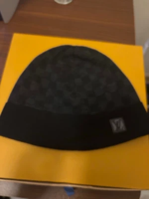 Svart Louis Vuitton mössa - Snygg svart mössa från Louis Vuitton med diskret rutmönster och vikbar kant. Märkets logga sitter på en liten patch framtill. Perfekt accessoar för dig som vill ha en exklusiv och stilren look. Materialet ser ut att vara mjukt och bekvämt.