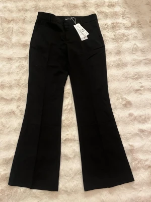 Lågmidjade Svarta utsvängda kostymbyxor Gina Tricot - Helt nya och aldrig använda! Svarta lågmidjade kostymbyxor från Gina Tricot med utsvängda ben och klassisk passform. Byxorna har hällor för skärp, fickor fram och en dekorativ bakficka. Perfekta för en stilren look. 