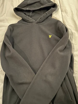 Svart hoodie från Lyle & Scott - Säljer en svart hoodie från Lyle & Scott med klassisk gul logga på bröstet. Hoodien har dragsko i huvan och är gjord i mjukt bomullsmaterial. Perfekt för en chill och stilren look. Står i storleken att den passar 14-15 åringar, jag skulle säga att det är storlek S.