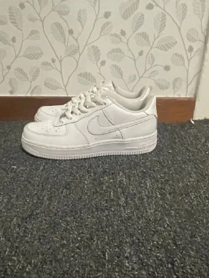 Nike Air Force 1 vita sneakers - Klassiska Nike Air Force 1 sneakers i helvitt läder med perforerad tå, kraftig sula och ikoniska swoosh-loggan på sidan. Skorna har snörning och diskret branding på hälen och plösen. Perfekta för dig som gillar stilrena och tidlösa sneakers och skorna kostar i nypris 1200 men säljer för 775