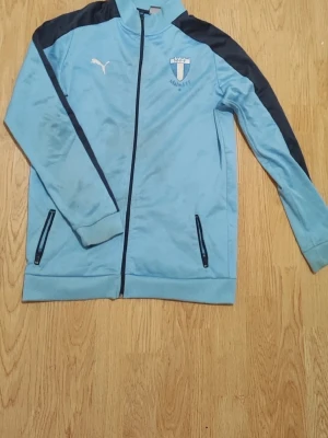 Ljusblå Malmö FF träningsjacka Puma - Ljusblå träningsjacka från Puma med Malmö FF-logga på bröstet och svarta detaljer längs axlar och ärmar. Jackan har hel dragkedja, ståkrage och två fickor med dragkedja. Perfekt för fotbollsträning eller sportig stil. Har några fläckar 