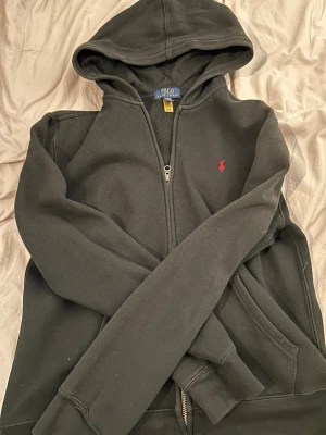 Svart hoodie från Polo Ralph Lauren - Klassisk svart hoodie från Polo Ralph Lauren med dragkedja framtill och huva. Tröjan har det ikoniska röda Polo-märket broderat på bröstet och mjukt bomullsmaterial. Perfekt för en avslappnad och stilren look. Storlek XL barnstorlek men passar vanlig S