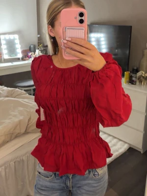 Röd smockad blus med volang - Trendig röd blus med smockad midja och volangkant. Blusen har rund halsringning och långa puffärmar med resår vid ärmslut. Perfekt för en färgstark look och passar till både jeans och kjol.